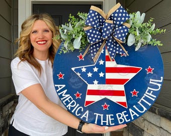 May include: A blue and red star-shaped door hanger with a white background. The star has red and white stripes and white stars. The text "America the Beautiful" is written in white around the star. The door hanger is decorated with a blue and white bow and greenery.