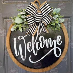 May include: A round wooden door sign with a black center and the word Welcome written in white script. The sign is decorated with a black and white striped bow and green foliage. The sign is suspended by a rope loop at the top of the frame.