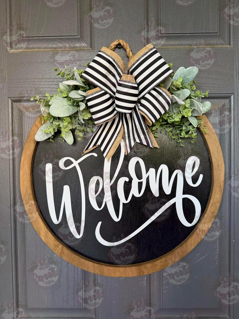 May include: A round wooden door sign with a black center and the word Welcome written in white script. The sign is decorated with a black and white striped bow and green foliage. The sign is suspended by a rope loop at the top of the frame.