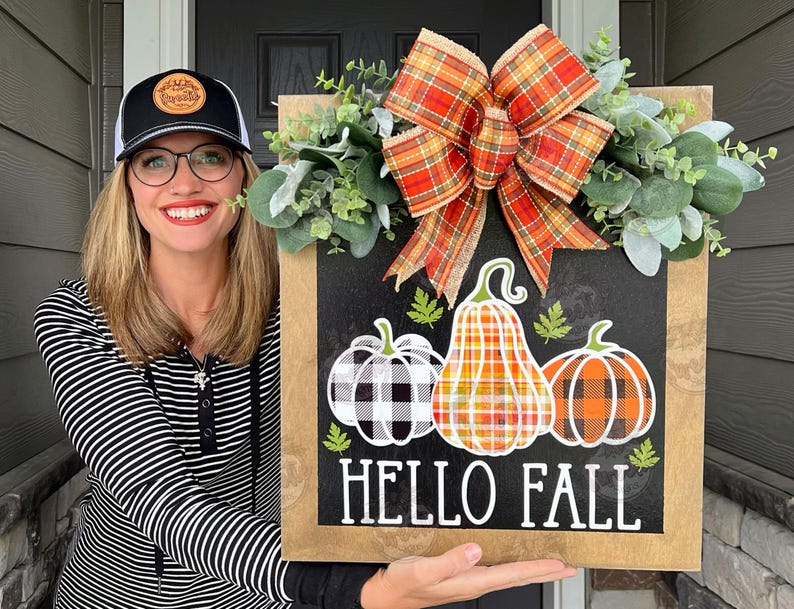 May include: A wooden sign with a black chalkboard background. The sign has a fall theme with a plaid bow, green leaves, and three pumpkins. The pumpkins are decorated with plaid and checkered patterns. The text on the sign reads 'Hello Fall'.
