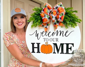 May include: A round, white wooden sign with the words "Welcome to our Home" in black script and block letters, featuring an orange pumpkin graphic. The sign is decorated with a green leafy garland and a bow with pumpkin and plaid patterns.