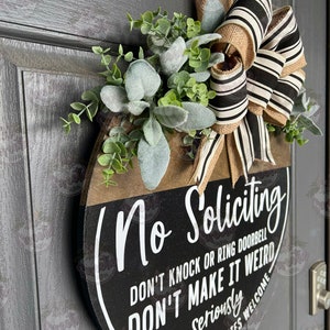 May include: A round wooden door sign with the words "No Soliciting" in white script. The sign has a black and brown color scheme, with a decorative bow and greenery. The text also reads "DON'T KNOCK OR RING DOORBELL DON'T MAKE IT WEIRD FRIENDS & DELIVERIES WELCOME".