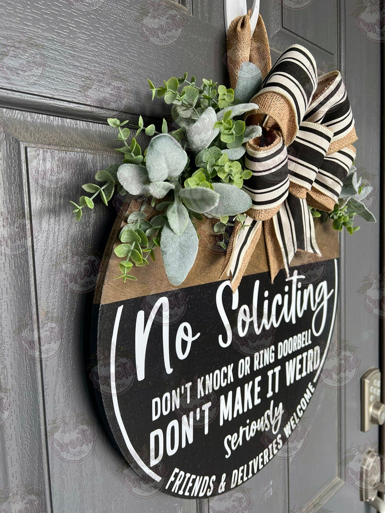 May include: A round wooden door sign with the words "No Soliciting" in white script. The sign has a black and brown color scheme, with a decorative bow and greenery. The text also reads "DON'T KNOCK OR RING DOORBELL DON'T MAKE IT WEIRD FRIENDS & DELIVERIES WELCOME".