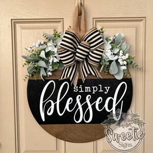 May include: A round wooden door sign with the words "simply blessed" in white script. The sign has a black and brown color scheme, adorned with a striped bow and faux greenery and flowers.
