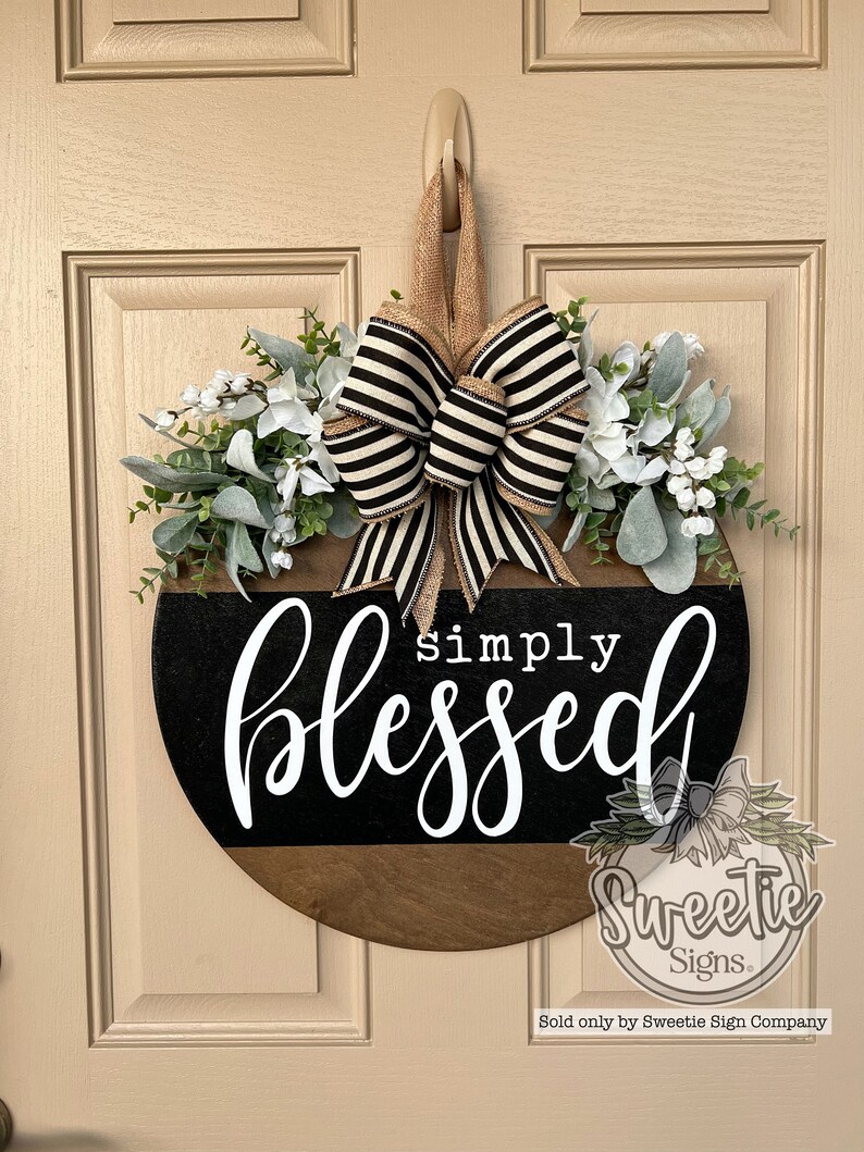 May include: A round wooden door sign with the words "simply blessed" in white script. The sign has a black and brown color scheme, adorned with a striped bow and faux greenery and flowers.