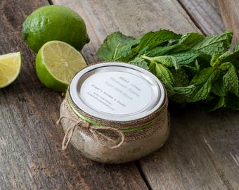 Puede incluir: Un tarro de vidrio con una tapa de arpillera marrón y una etiqueta blanca que dice "Mint Lime Homemade Oatmeal Sugar Scrub & Bath Salts". El tarro está rodeado de hojas de menta verde frescas y limas.