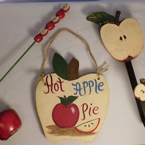 May include: Three wooden apple-shaped decorations. One is a sign that says "Hot Apple Pie" with a red apple and a slice of apple painted on it. Another is a red apple with a green stem and leaves. The third is a hook with a small apple on the end.