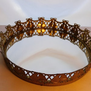 May include: A round gold-toned decorative tray with a mirrored surface. The tray has a decorative border with a repeating pattern of bells.