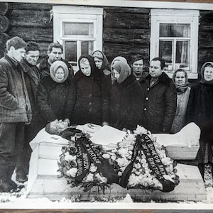May include: A black and white photograph of a group of people standing around a casket covered in flowers. The casket is on a wooden platform. The people are dressed in dark clothing and have somber expressions on their faces. The photo was taken in front of a wooden building with windows.