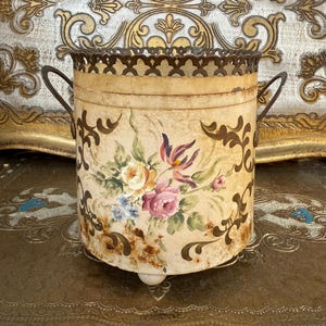 Może przedstawiać: Kremowy metalowy pojemnik z kwiatowym i zwijanym wzorem. Pojemnik ma ozdobną krawędź i uchwyty. Malowane kwiaty obejmują różowe, żółte i niebieskie kwiaty. Pojemnik ma około 15 cm wysokości.