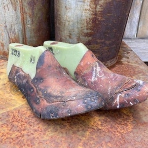 May include: Two brown leather shoe lasts with green and white markings. The lasts are sitting on a rusty metal surface. The lasts are marked with the numbers 99 and 3273.