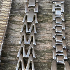 Może przedstawiać: Trzy r&oacute;żne style metalowych pask&oacute;w do zegark&oacute;w. Paski są srebrne i mają r&oacute;żne wzory ogniw. Paski są ułożone poziomo na drewnianej powierzchni.