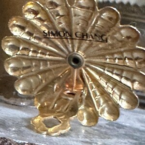 K&ouml;nnte beinhalten: Ein goldfarbenes, dekoratives Metallobjekt mit einem f&auml;cherf&ouml;rmigen Design und dem Text "SIMON CHANG" in der Mitte. Das Objekt hat ein kleines Loch in der Mitte und eine Schlaufe unten.