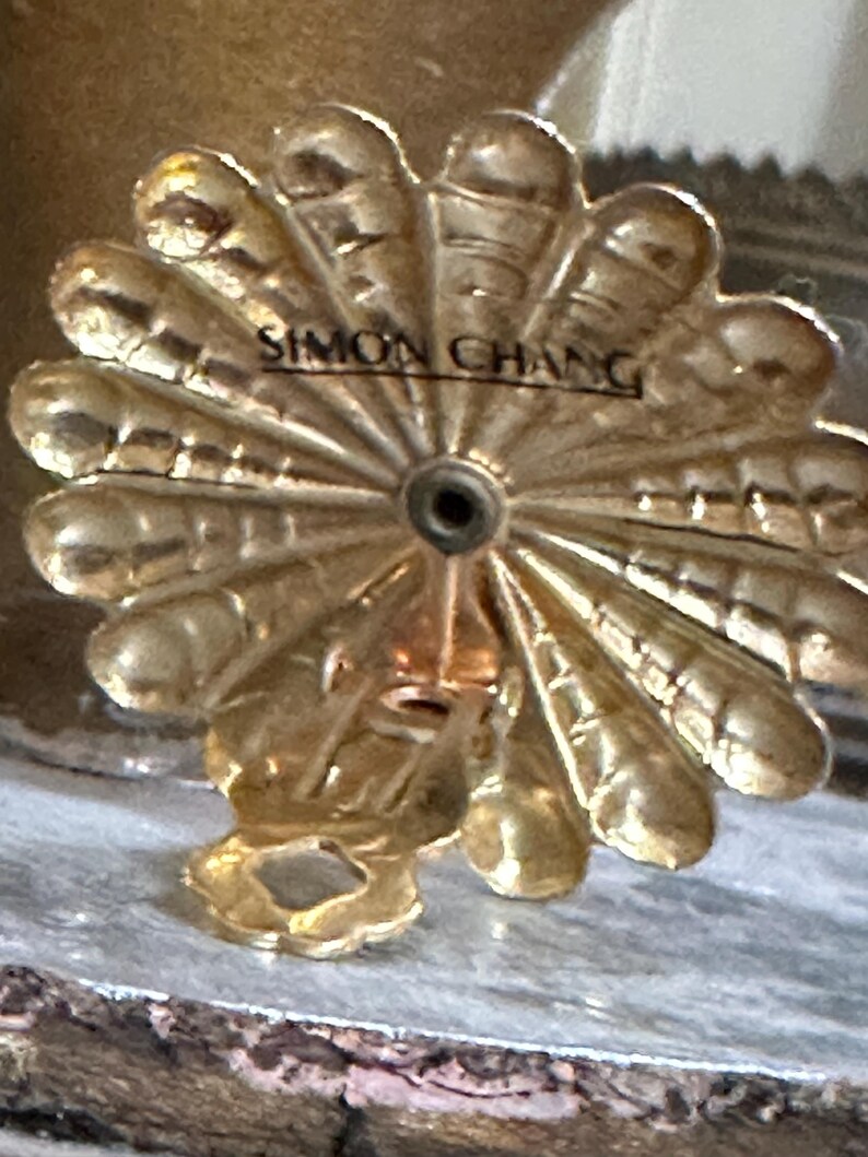 K&ouml;nnte beinhalten: Ein goldfarbenes, dekoratives Metallobjekt mit einem f&auml;cherf&ouml;rmigen Design und dem Text "SIMON CHANG" in der Mitte. Das Objekt hat ein kleines Loch in der Mitte und eine Schlaufe unten.