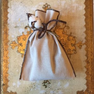 May include: A small, off-white fabric pouch with a brown drawstring closure. The pouch is tied with a brown cord and has a small bunch of white cotton on top.