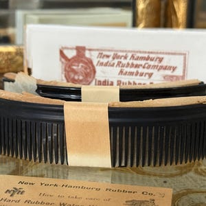 Puede incluir: Primer plano de un peine negro con dientes finos, envuelto en papel marrón. El peine está frente a una caja blanca con el texto "New York Hamburg India Rubber Company Hamburg India Rubber". Una pequeña tarjeta con texto está en primer plano.