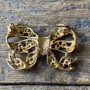 May include: Gold-toned bow brooch with a delicate, intricate design. The bow is made of metal with a textured finish and features a pin on the back for attaching to clothing.