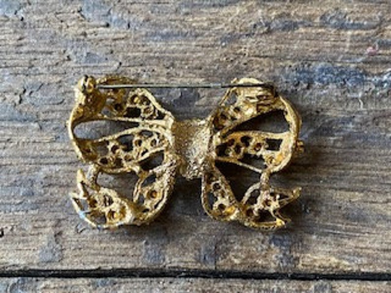 May include: Gold-toned bow brooch with a delicate, intricate design. The bow is made of metal with a textured finish and features a pin on the back for attaching to clothing.
