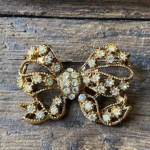 May include: A gold-toned brooch in the shape of a bow, with clear rhinestones covering the entire surface.