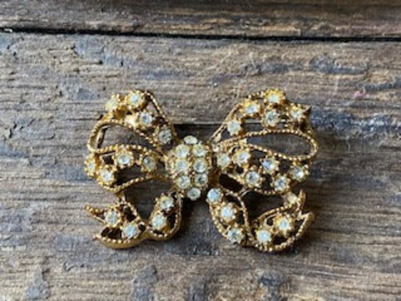 May include: A gold-toned brooch in the shape of a bow, with clear rhinestones covering the entire surface.