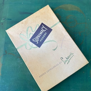 Puede incluir: Una caja vintage para medias, con el texto "Supremacy Hosiery" en un rectángulo morado con borde blanco. La caja es marrón y tiene el texto "Sheer Loveliness by Simpsons" impreso en la parte delantera.