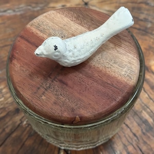 Puede incluir: Un tarro de vidrio decorativo con tapa de madera. Una figura de pájaro blanco descansa sobre la tapa. El tarro tiene un cuerpo de vidrio transparente y texturizado. La tapa de madera es redonda y de color marrón rojizo. La figura del pájaro es blanca con detalles negros.