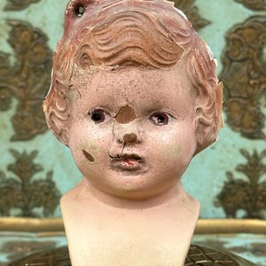 May include: A vintage doll head made of painted plaster with a cracked surface. The doll has brown hair, blue eyes, and a painted mouth. The head is missing part of the back of the head and the top of the head.
