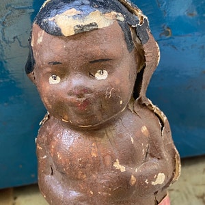 May include: Vintage doll with a brown face, black hair, and white eyes. The doll is made of papier-mâché and is in a worn condition.
