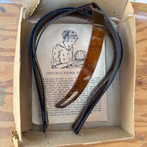 Könnte beinhalten: Vintage Haarstyling-Set in einem Pappkarton. Das Set enthält einen braunen und schwarzen Kunststoff-Haarkamm und ein gedrucktes Anleitungsblatt mit einer Zeichnung einer Frau, die sich die Haare stylt. Die Box ist abgenutzt.