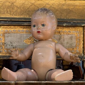 Puede incluir: Muñeca de bebé vintage con cara pintada, ojos azules y labios rojos. La muñeca está sentada con los brazos extendidos. La piel de la muñeca es de color bronceado claro y tiene cabello moldeado. Un juguete clásico.