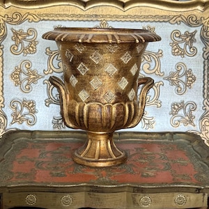 Op de afbeelding: Een goudkleurige decoratieve urn met een voetstuk en twee handvatten. De urn heeft een textuuroppervlak met ruitvormige patronen. Hij staat op een rood en goud dienblad met sierlijke details, tegen een lichtblauwe achtergrond.