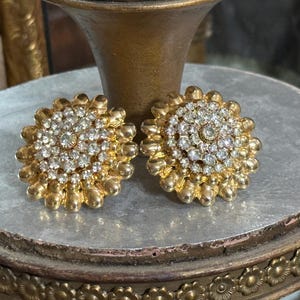 K&ouml;nnte beinhalten: Ein Paar goldfarbene Ohrclips mit Blumenmuster. Die Ohrringe sind mit klaren Strasssteinen verziert.