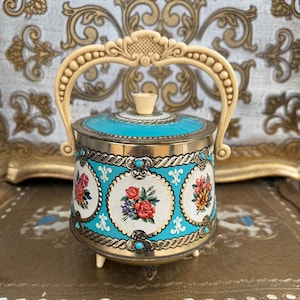 May include: Vintage metal container with a turquoise lid and floral designs. The container has a cream handle and feet, and is adorned with gold and turquoise accents. The floral designs include roses and other flowers.