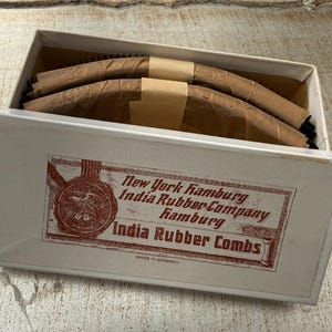 Puede incluir: Una caja blanca con una etiqueta roja y negra que dice "New York Hamburg India Rubber Company Hamburg India Rubber Combs Made in Germany". La caja contiene varios peines curvos marrones envueltos en papel marrón.