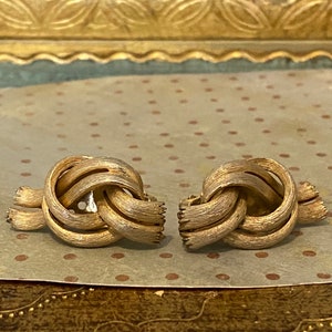 Może przedstawiać: Para złotych kolczyków w kształcie węzła. Kolczyki wykonane są z fakturowanego metalu i mają prosty, elegancki design.
