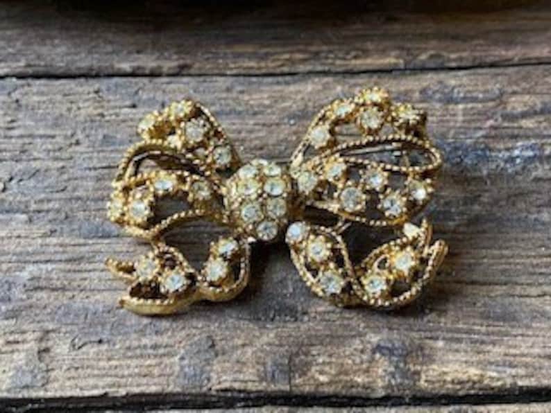 May include: A gold-toned bow brooch with clear rhinestones. The bow is made of metal and has a detailed design. The rhinestones are set in a cluster in the center of the bow.
