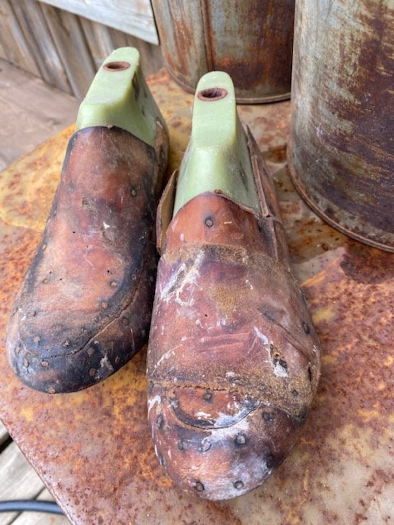 May include: Two brown leather shoe lasts with green plastic inserts. The lasts are sitting on a rusty metal surface.