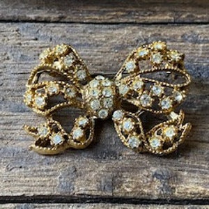May include: A gold-toned brooch in the shape of a bow, adorned with clear rhinestones.