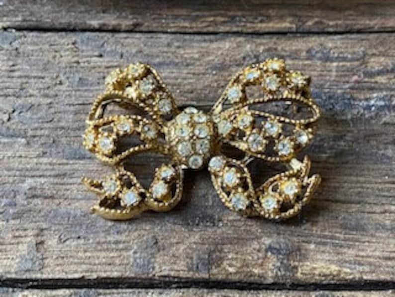May include: A gold-toned brooch in the shape of a bow, adorned with clear rhinestones.