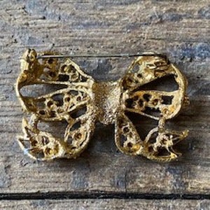 May include: A gold-toned bow brooch with clear rhinestones. The bow is detailed with a filigree design and has a pin back.