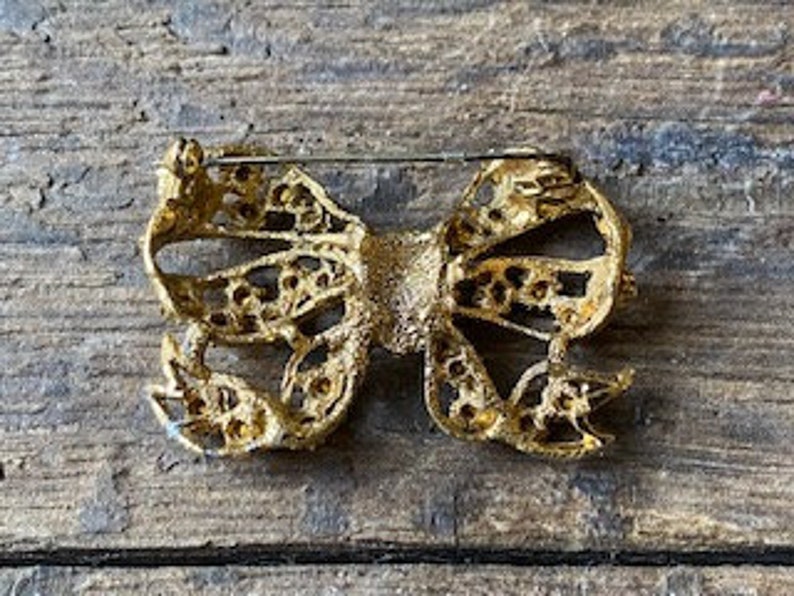 May include: A gold-toned bow brooch with clear rhinestones. The bow is detailed with a filigree design and has a pin back.