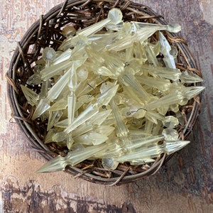 Op de afbeelding: Een verzameling vintage, lichtgele kroonkristallen in een geweven bruine mand. De kristallen hebben een puntige, ijspegelachtige vorm met facetdetails. De mand staat op een getextureerd, lichtbruin houten oppervlak.