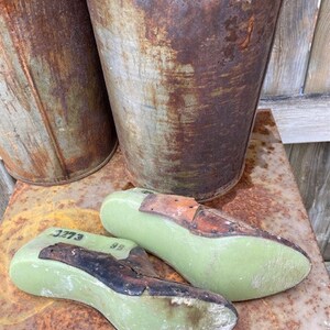 May include: Two green shoe lasts with brown leather tops. The lasts are numbered 3773 and 98. The lasts are sitting on a rusty metal surface.