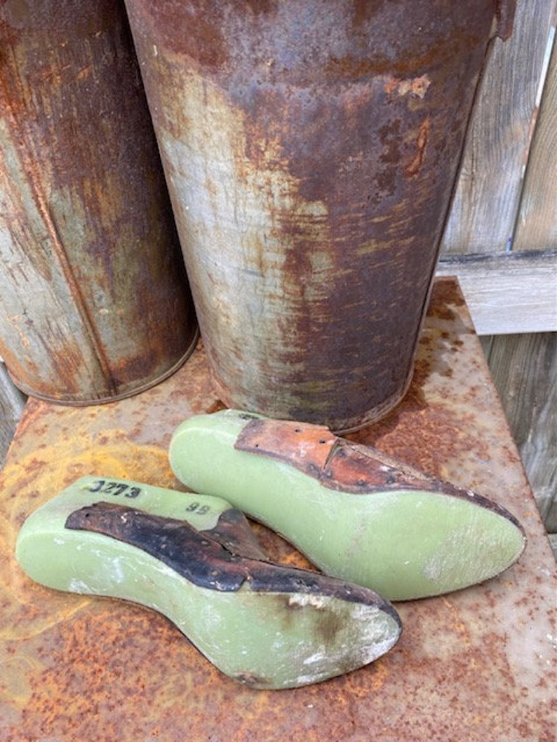 May include: Two green shoe lasts with brown leather tops. The lasts are numbered 3773 and 98. The lasts are sitting on a rusty metal surface.