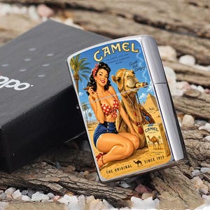 May include: A silver-colored Zippo lighter with a vintage Camel cigarettes graphic. The graphic features a woman in a polka dot bikini and shorts, a camel smoking a cigar, and the text "CAMEL" and "SINCE 1913". The lighter is in a black box.