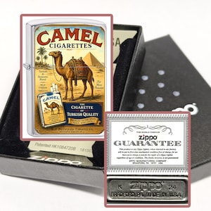 Puede incluir: Un encendedor Zippo plateado con un dise&ntilde;o de Camel Cigarettes. La obra de arte presenta un camello, pir&aacute;mides y texto. El encendedor est&aacute; en una caja negra con una tarjeta de garant&iacute;a y una base de metal con el logotipo de Zippo.