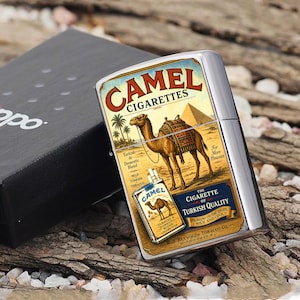 Puede incluir: Un encendedor Camel Cigarettes plateado con un dise&ntilde;o vintage. El encendedor presenta una ilustraci&oacute;n de camello con una pir&aacute;mide y palmeras. La caja es negra con la palabra "Zippo". El encendedor est&aacute; sobre un lecho de virutas de madera.