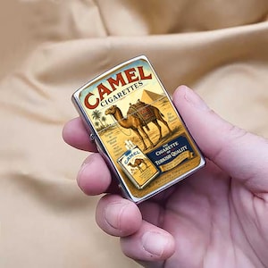 Puede incluir: Un encendedor Camel Cigarettes de metal plateado. El encendedor presenta un dise&ntilde;o vintage con un camello, palmeras y una pir&aacute;mide. El texto "Camel Cigarettes" se muestra en la parte superior.