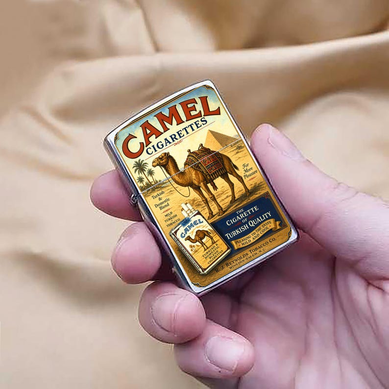 Puede incluir: Un encendedor Camel Cigarettes de metal plateado. El encendedor presenta un dise&ntilde;o vintage con un camello, palmeras y una pir&aacute;mide. El texto "Camel Cigarettes" se muestra en la parte superior.