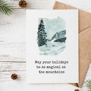 Peut inclure: Carte de vœux de Noël avec une scène de montagne à l'aquarelle et le texte "May your holidays be as magical as the mountains". La carte est accompagnée d'une enveloppe marron, de pommes de pin et de branches de conifères.